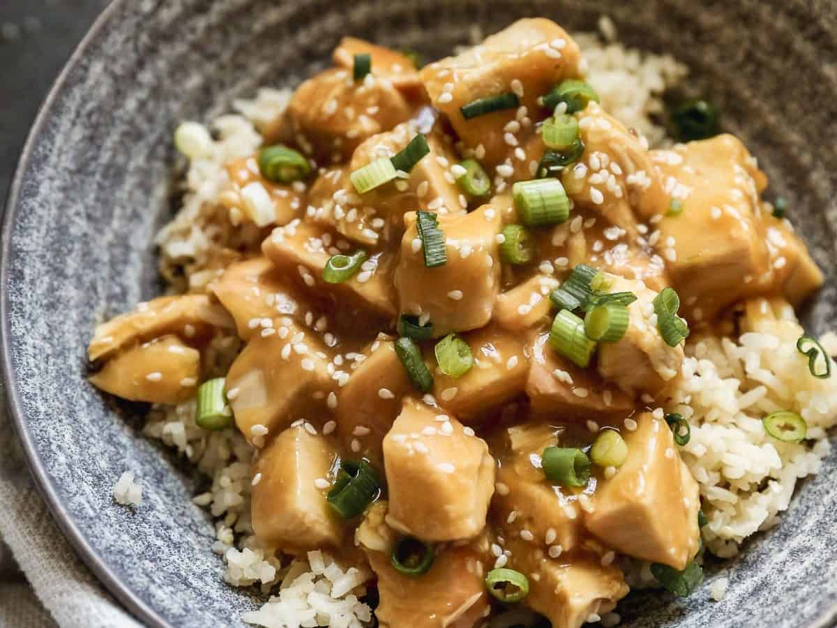 A bowl of rice topped with slow cooker orange chicken, garnished with chopped green onions and sesame seeds. The dish is served in a gray ceramic bowl on a dark surface.