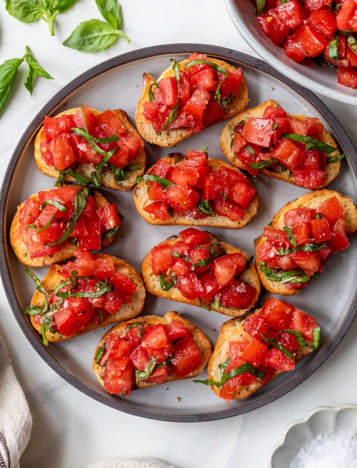 A round plate holds several pieces of bruschetta topped with diced tomatoes and chopped basil, arranged neatly on toasted bread slices. Fresh basil leaves are scattered nearby on a white surface.