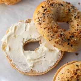 An everything cottage cheese bagel sliced in half, with one half spread with cream cheese and the other half resting nearby on parchment paper. Two more whole everything bagels are partially visible.