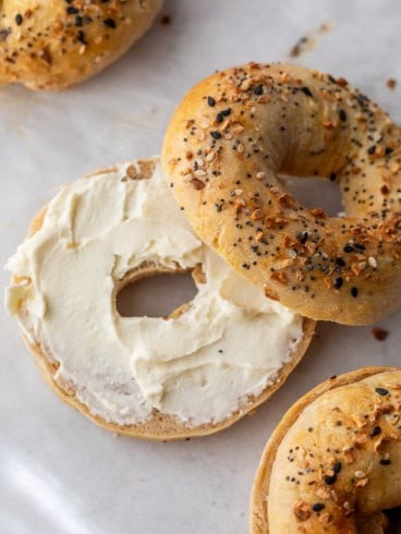An everything cottage cheese bagel sliced in half, with one half spread with cream cheese and the other half resting nearby on parchment paper. Two more whole everything bagels are partially visible.