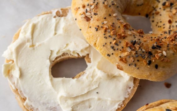 An everything cottage cheese bagel sliced in half, with one half spread with cream cheese and the other half resting nearby on parchment paper. Two more whole everything bagels are partially visible.