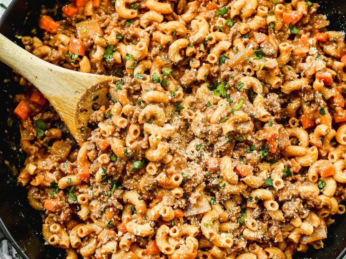 A large black skillet filled with ground beef pasta, topped with grated cheese and chopped parsley. A wooden spoon rests in the pasta, and a striped towel sits nearby.