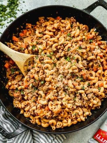 A large black skillet filled with ground beef pasta, topped with grated cheese and chopped parsley. A wooden spoon rests in the pasta, and a striped towel sits nearby.