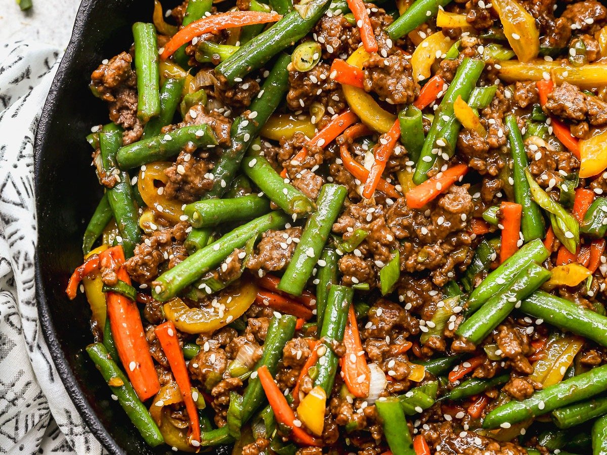 A skillet filled with ground beef stir fry with green beans, red and yellow bell peppers, garnished with sesame seeds and sliced green onions. A patterned cloth is partially visible beneath the skillet.