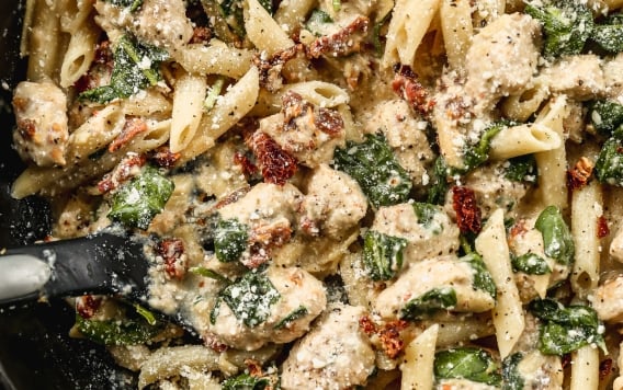 A skillet filled with creamy high protein pasta, chunks of chicken, spinach, and sun-dried tomatoes, topped with grated Parmesan cheese.