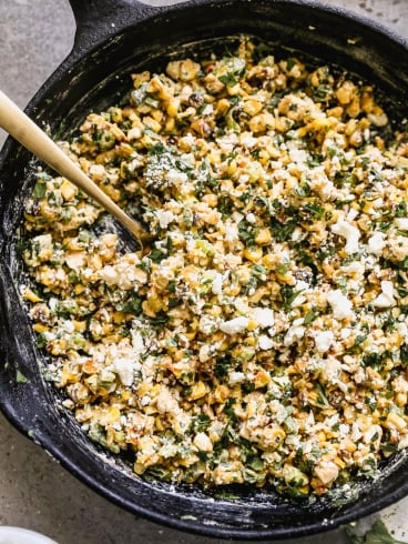 A cast iron skillet filled with Mexican street corn salad, with a wooden spoon resting in the pan.