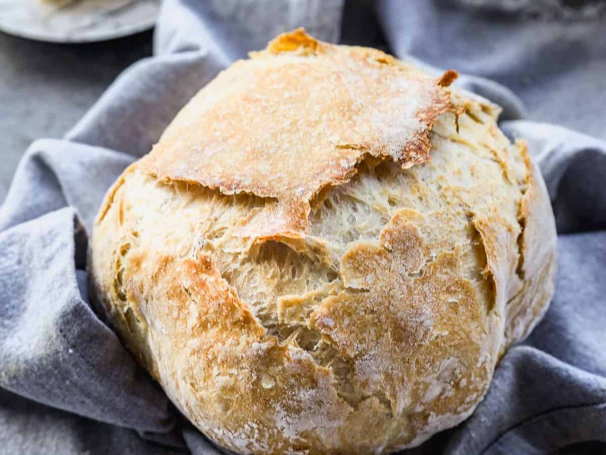 A round loaf of rustic, crusty no-knead bread sits on a gray cloth, with a plate of butter and a butter knife in the background on a gray surface.