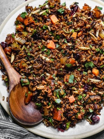 A plate of wild rice pilaf mixed with chopped carrots, celery, herbs, nuts, and dried cranberries, with a wooden serving spoon resting on the edge of the plate.