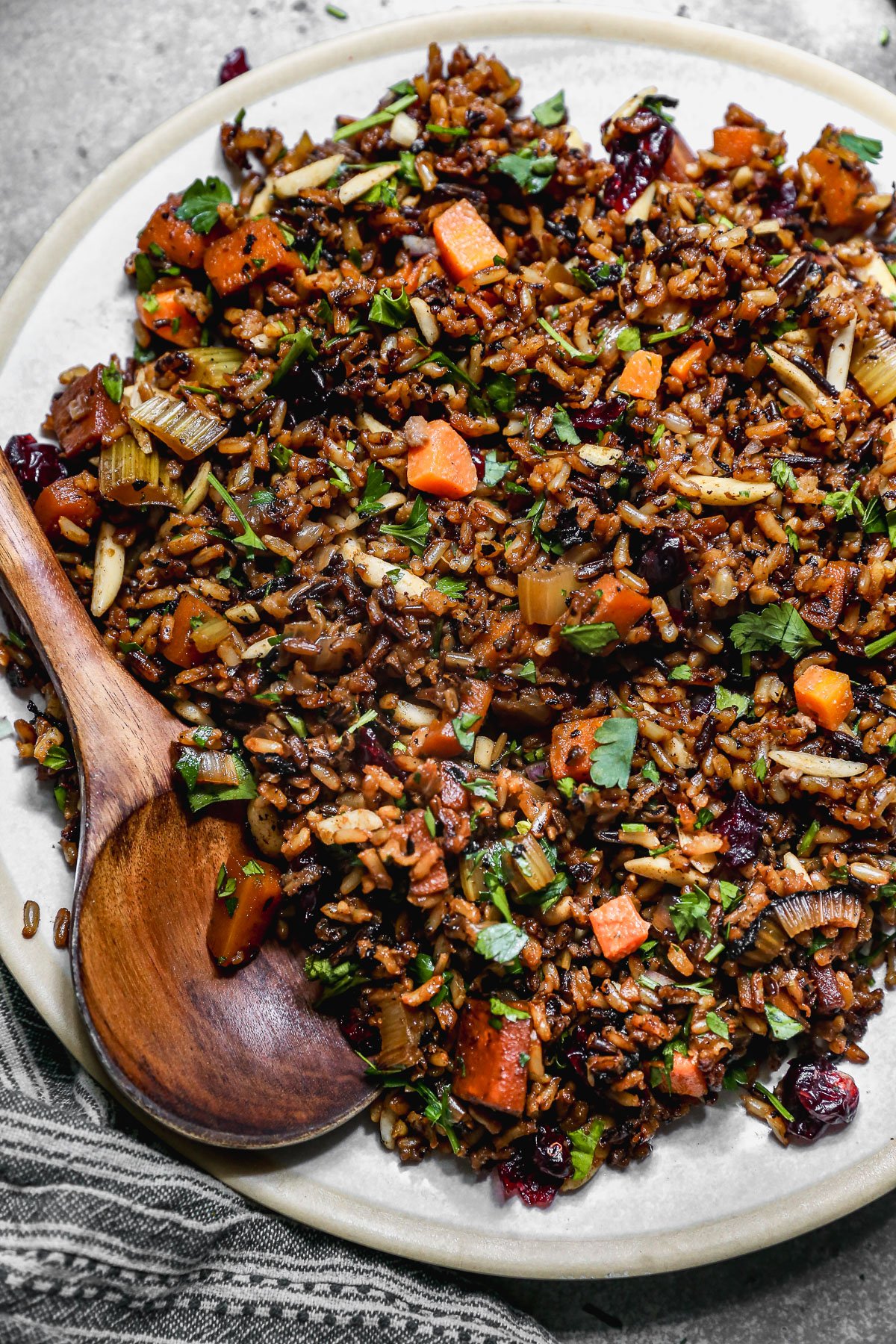 A plate of wild rice pilaf mixed with chopped carrots, celery, herbs, nuts, and dried cranberries, with a wooden serving spoon resting on the edge of the plate.