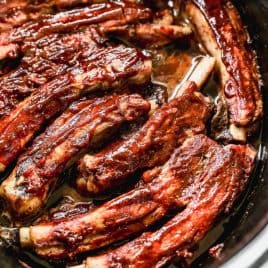 Barbecue pork ribs coated in sauce, slow-cooked in a crockpot, with meat glistening and slightly caramelized, arranged closely together in a dark, oval-shaped cooker.