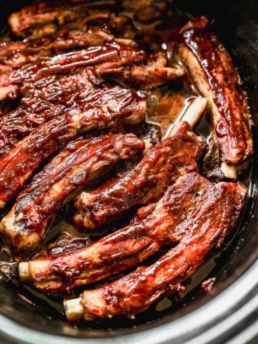 Barbecue pork ribs coated in sauce, slow-cooked in a crockpot, with meat glistening and slightly caramelized, arranged closely together in a dark, oval-shaped cooker.