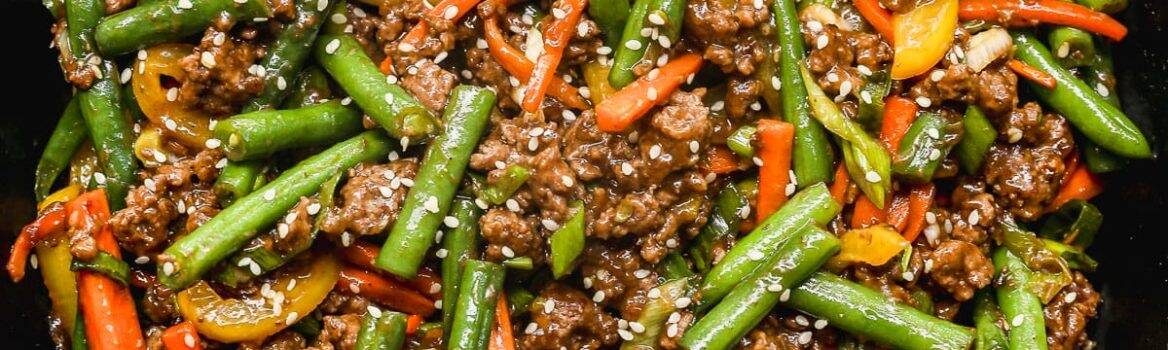 A skillet filled with stir-fried ground beef, green beans, red and yellow bell peppers, and carrots, garnished with sesame seeds and chopped green onions.
