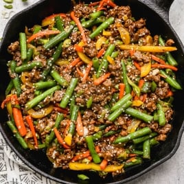 A skillet filled with stir-fried ground beef, green beans, red and yellow bell peppers, and carrots, garnished with sesame seeds and chopped green onions.