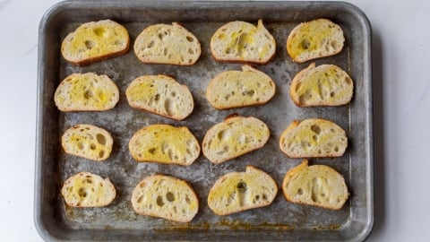 A metal baking tray holds fifteen slices of rustic bread, each drizzled with olive oil and spaced evenly on the tray, ready for toasting. The tray sits on a white surface.