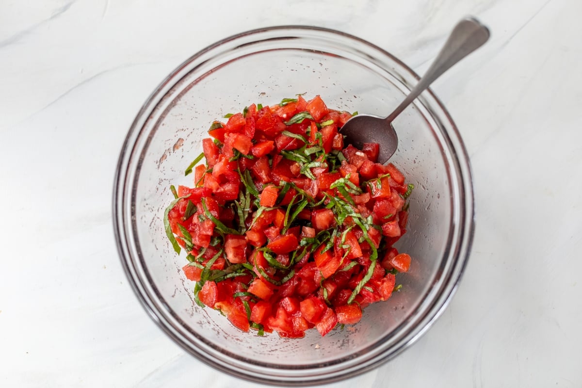 A glass bowl filled with a mixture of diced tomatoes and chopped fresh basil for bruschetta, with a spoon resting inside, on a white surface.