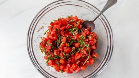 A glass bowl filled with a mixture of diced tomatoes and chopped fresh basil for bruschetta, with a spoon resting inside, on a white surface.