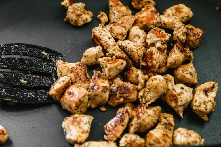 Chunks of seasoned, cooked chicken breast are being sautéed in a pan, with a black spatula visible on the left side. The chicken pieces have a golden-brown sear on the outside.