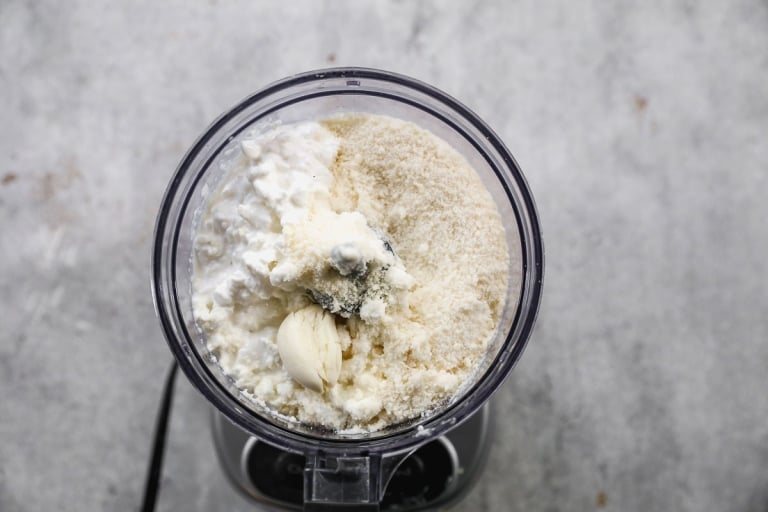 A top-down view of a food processor filled with cottage cheese, grated parmesan cheese, and a clove of garlic on a gray countertop.