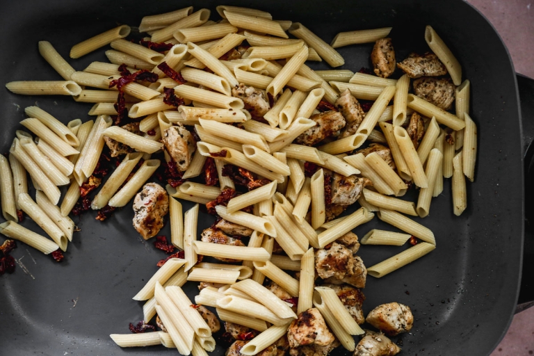 Cooked penne high protein pasta, pieces of seasoned chicken, and small bits of sun-dried tomato are in a skillet, partially mixed, ready for further cooking or serving.