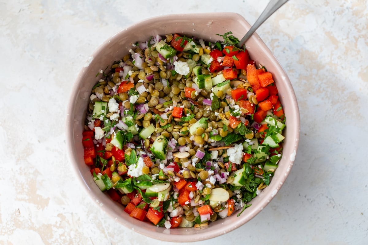 A pink bowl filled with a colorful salad containing chopped red bell peppers, cucumbers, red onions, lentils, feta cheese, and fresh herbs, with a spoon resting inside the bowl.
