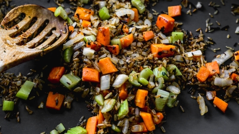 A close-up of a wooden spoon resting on a mix of cooked wild rice, diced carrots, celery, and onions on a dark surface.