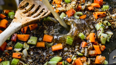 Chopped carrots, celery, onion, and wild rice cooking in a pan with broth being poured in, stirred with a wooden spoon.