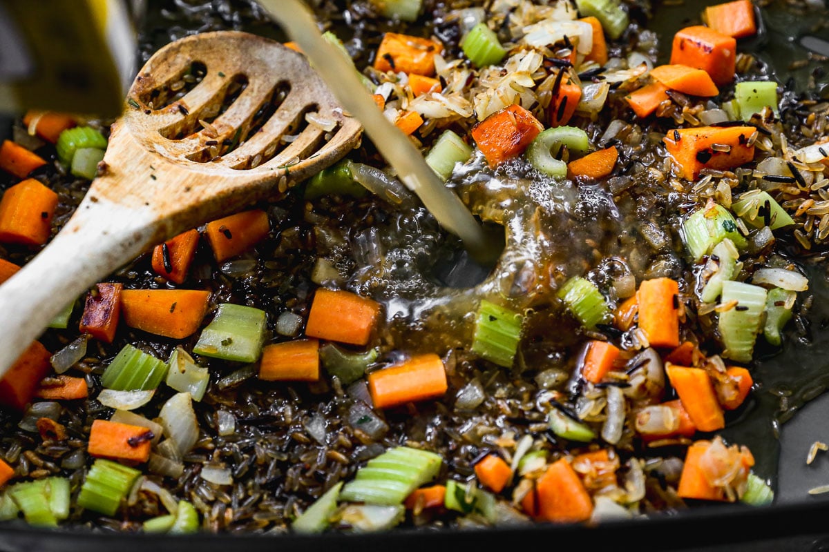 Chopped carrots, celery, onion, and wild rice cooking in a pan with broth being poured in, stirred with a wooden spoon.