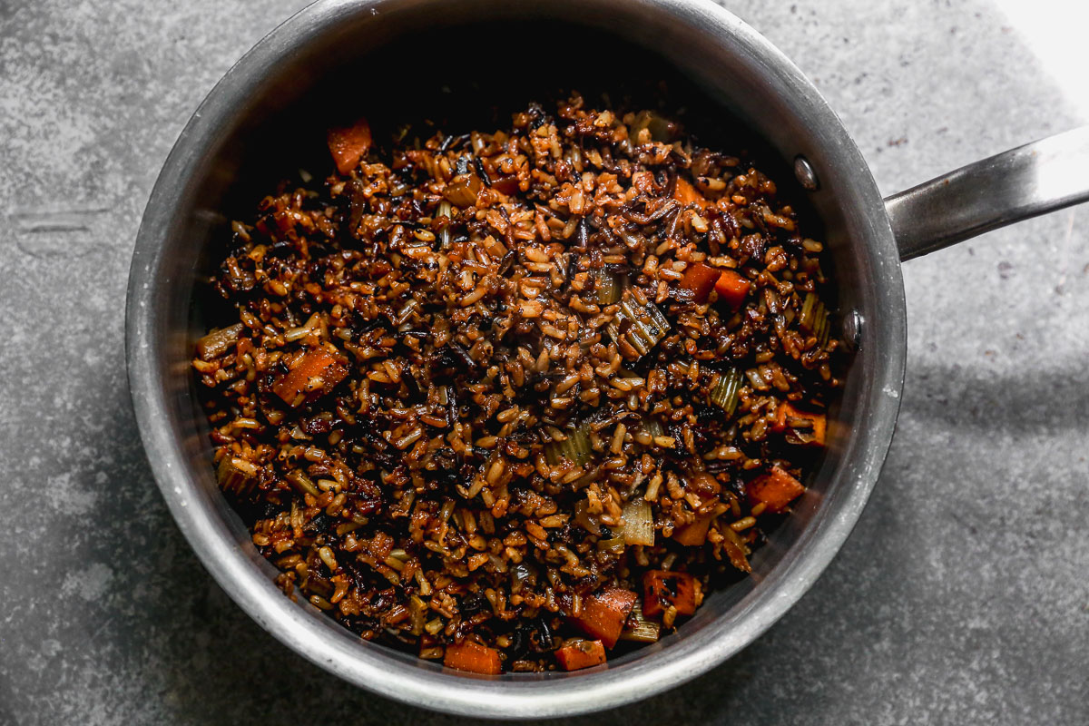 A metal pot filled with cooked wild rice pilaf mixed with chopped vegetables, including carrots and celery, sits on a gray surface. The rice and vegetables appear well-mixed and slightly browned.
