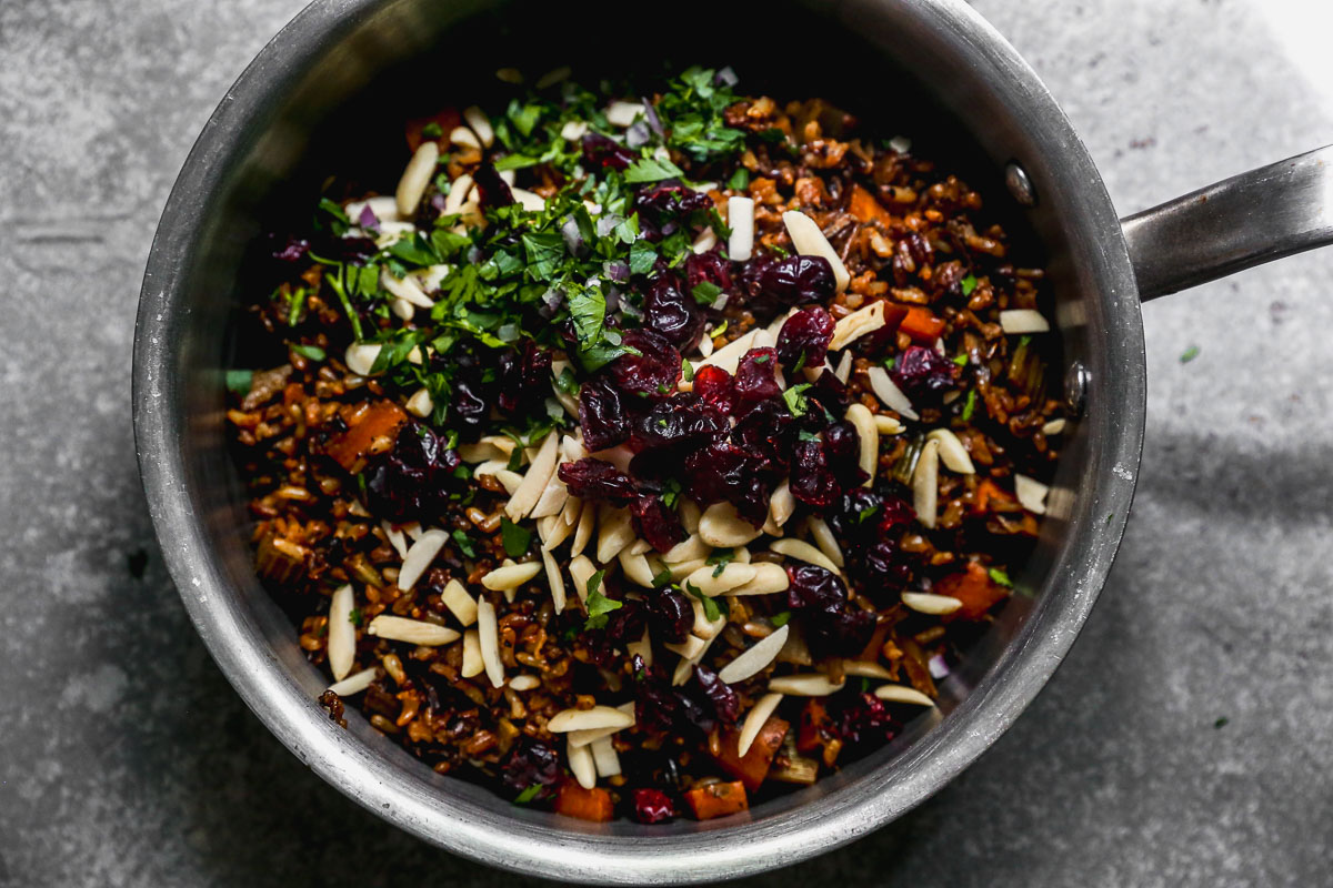 A overhead view of a saucepan filled with a mixture of cooked wild rice, diced vegetables, slivered almonds, dried cranberries, and chopped fresh herbs on a gray surface.