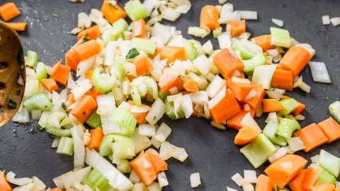 Chopped onions, carrots, and celery pieces are being sautéed in a pan, with a wooden spoon partially visible on the side.