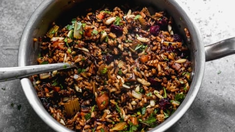 A metal pan filled with wild rice pilaf mixed with chopped vegetables, herbs, nuts, and dried cranberries. A metal spoon rests inside the pan on a gray countertop.