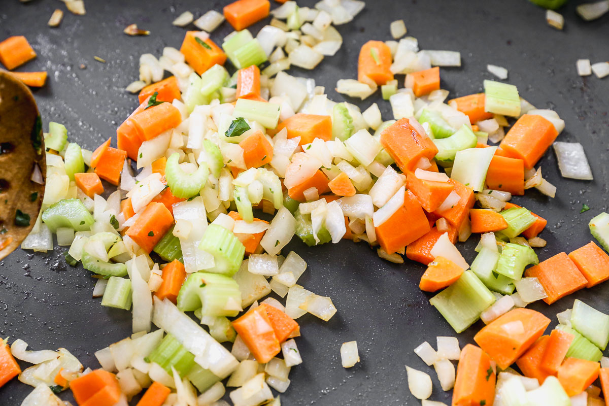 Chopped onions, carrots, and celery pieces are being sautéed in a pan, with a wooden spoon partially visible on the side.