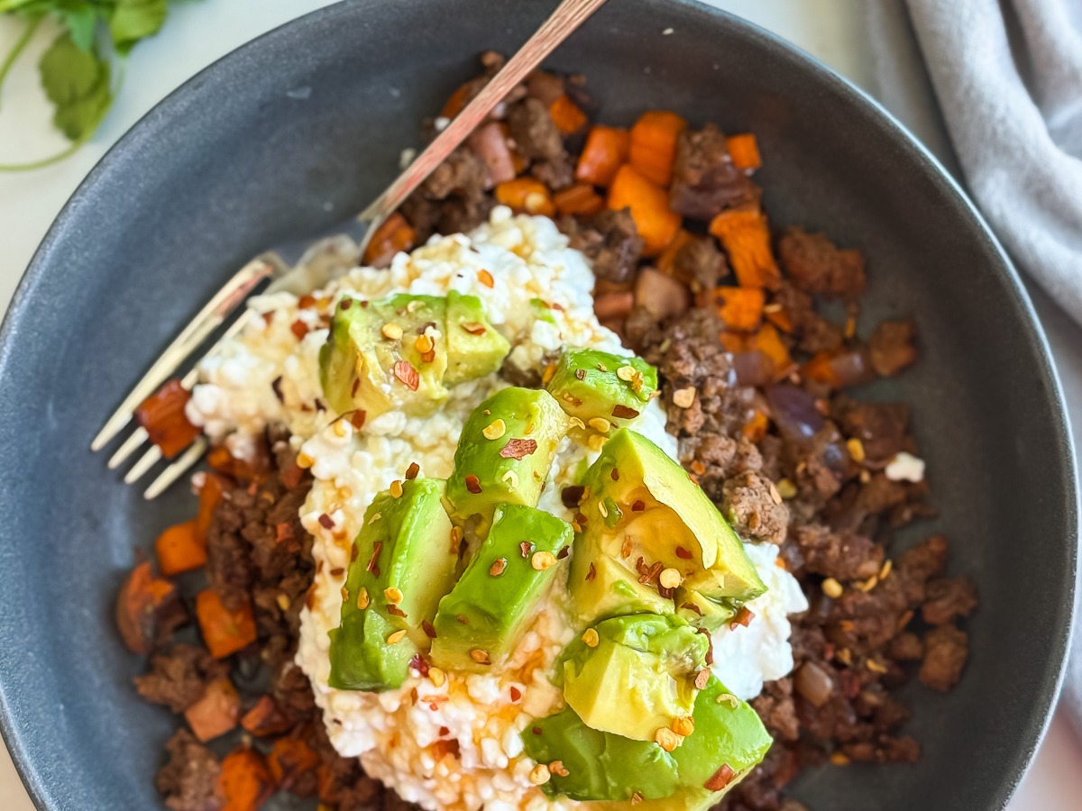 A ground beef bowl with ground beef, diced sweet potatoes, and onions, topped with cottage cheese, avocado chunks, and red pepper flakes. A silver fork rests in the bowl. Avocado, cilantro, and a gray napkin are nearby.