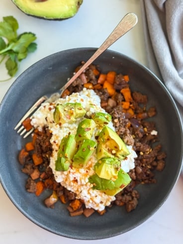 A ground beef bowl with ground beef, diced sweet potatoes, and onions, topped with cottage cheese, avocado chunks, and red pepper flakes. A silver fork rests in the bowl. Avocado, cilantro, and a gray napkin are nearby.