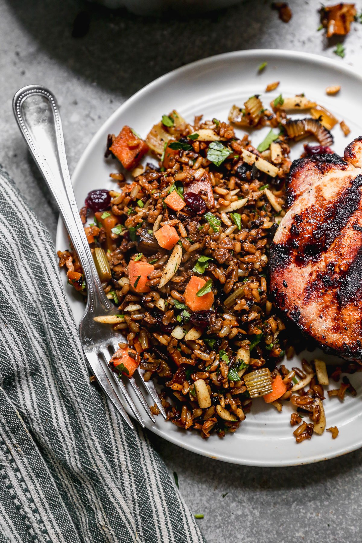 A plate with grilled chicken and a serving of wild rice pilaf mixed with carrots, celery, almonds, and herbs. A fork rests on the plate next to a green and white striped napkin.