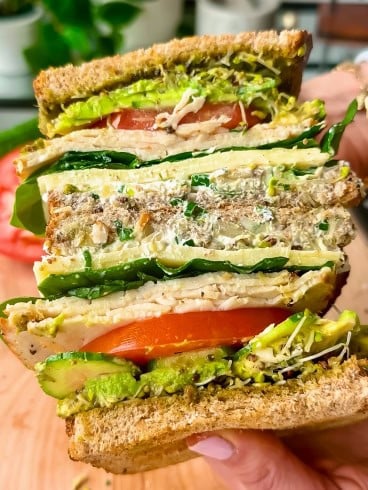 A close-up of a hand holding a thick sandwich filled with layers of tomatoes, lettuce, sprouts, avocado, cheese, and whole grain bread, with visible seeds and a colorful, fresh appearance.