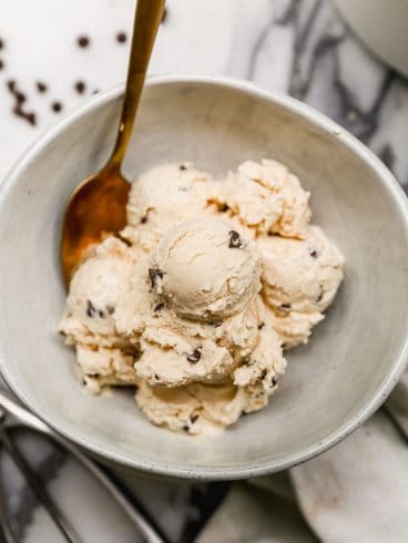 A bowl of creamy cottage cheese ice cream with chocolate chips, served with a gold spoon on a marble surface.