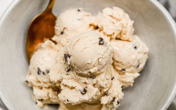 A bowl of creamy cottage cheese ice cream with chocolate chips, served with a gold spoon on a marble surface.