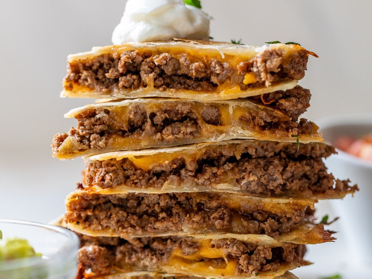 A stack of cheese and beef quesadillas cut into wedges, topped with a dollop of sour cream and garnished with cilantro. A bowl of guacamole and some herbs are visible in the foreground.