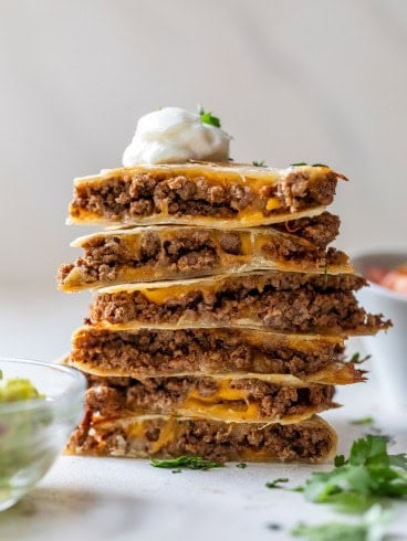 A stack of cheese and beef quesadillas cut into wedges, topped with a dollop of sour cream and garnished with cilantro. A bowl of guacamole and some herbs are visible in the foreground.