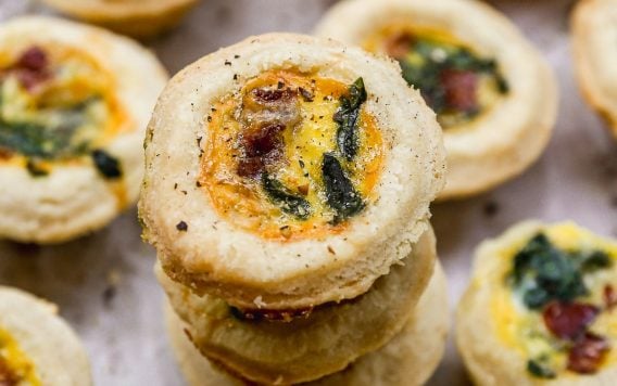 Mini quiche bites with golden crusts, filled with egg, herbs, and bits of bacon, arranged on a light surface.