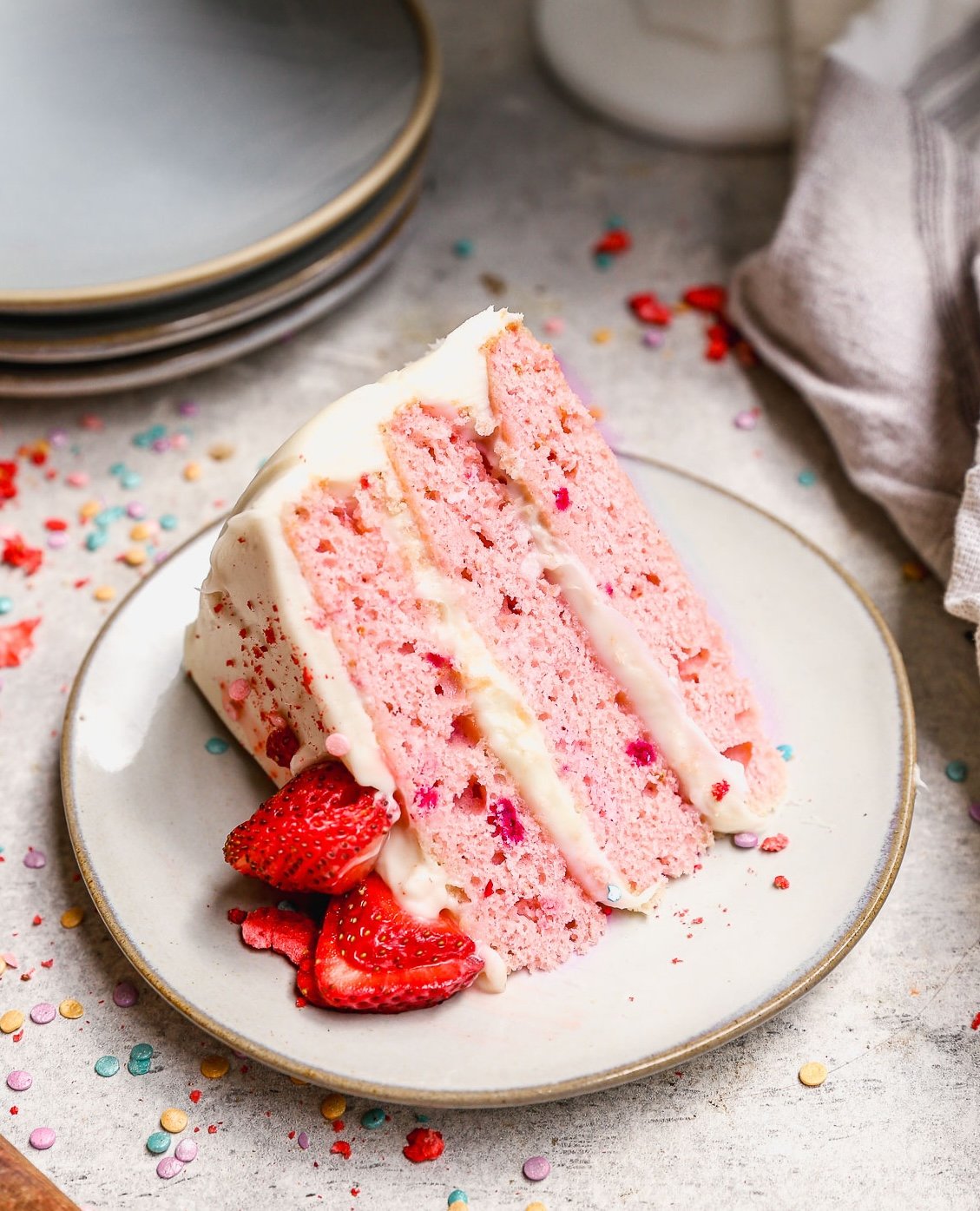 A slice of pink cake with three layers of white frosting, garnished with fresh sliced strawberries, sits on a plate. Colorful sprinkles are scattered around on the light-colored table.