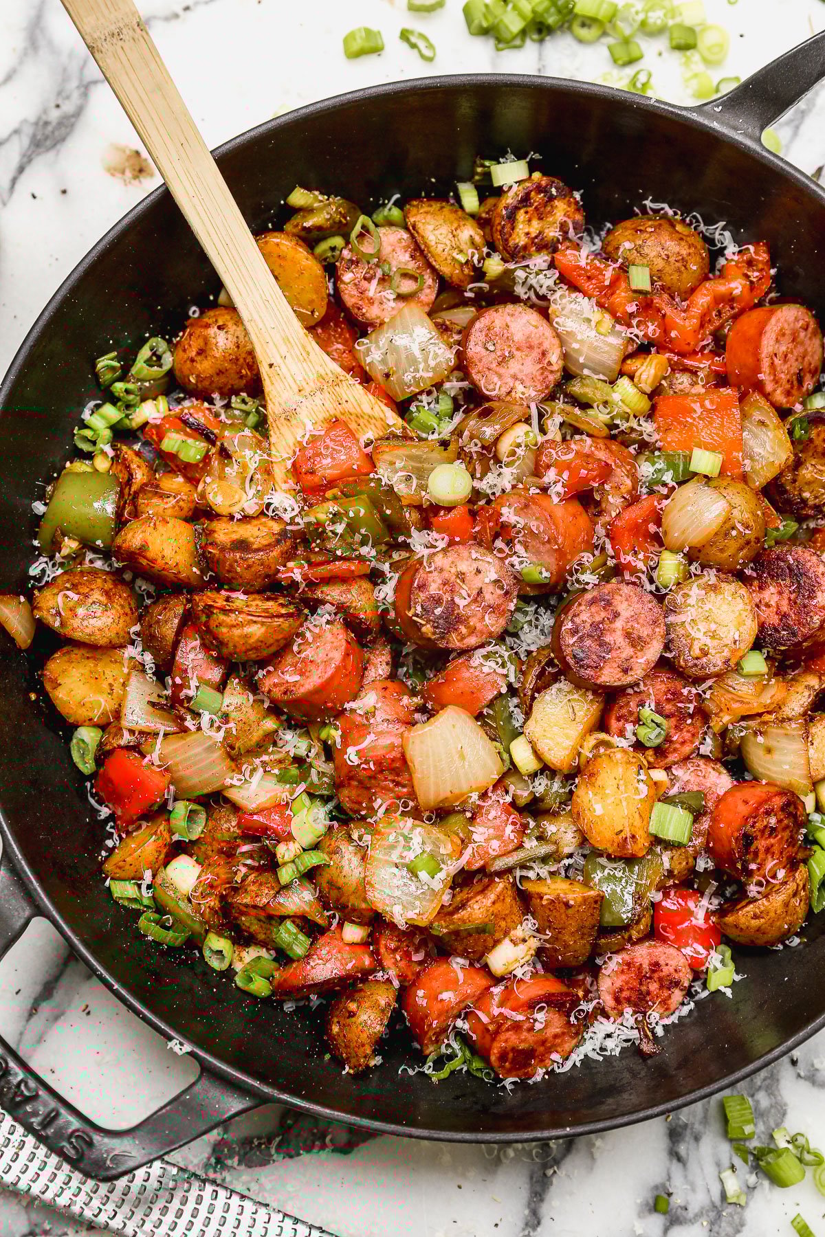 A skillet filled with sautéed sausage slices, diced potatoes, onions, bell peppers, and tomatoes, garnished with chopped green onions and grated cheese, with a wooden spoon resting inside.