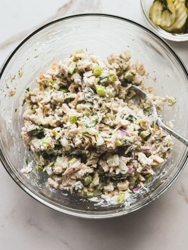A glass bowl filled with creamy chicken salad mixed with chopped celery, red onion, and dill pickles