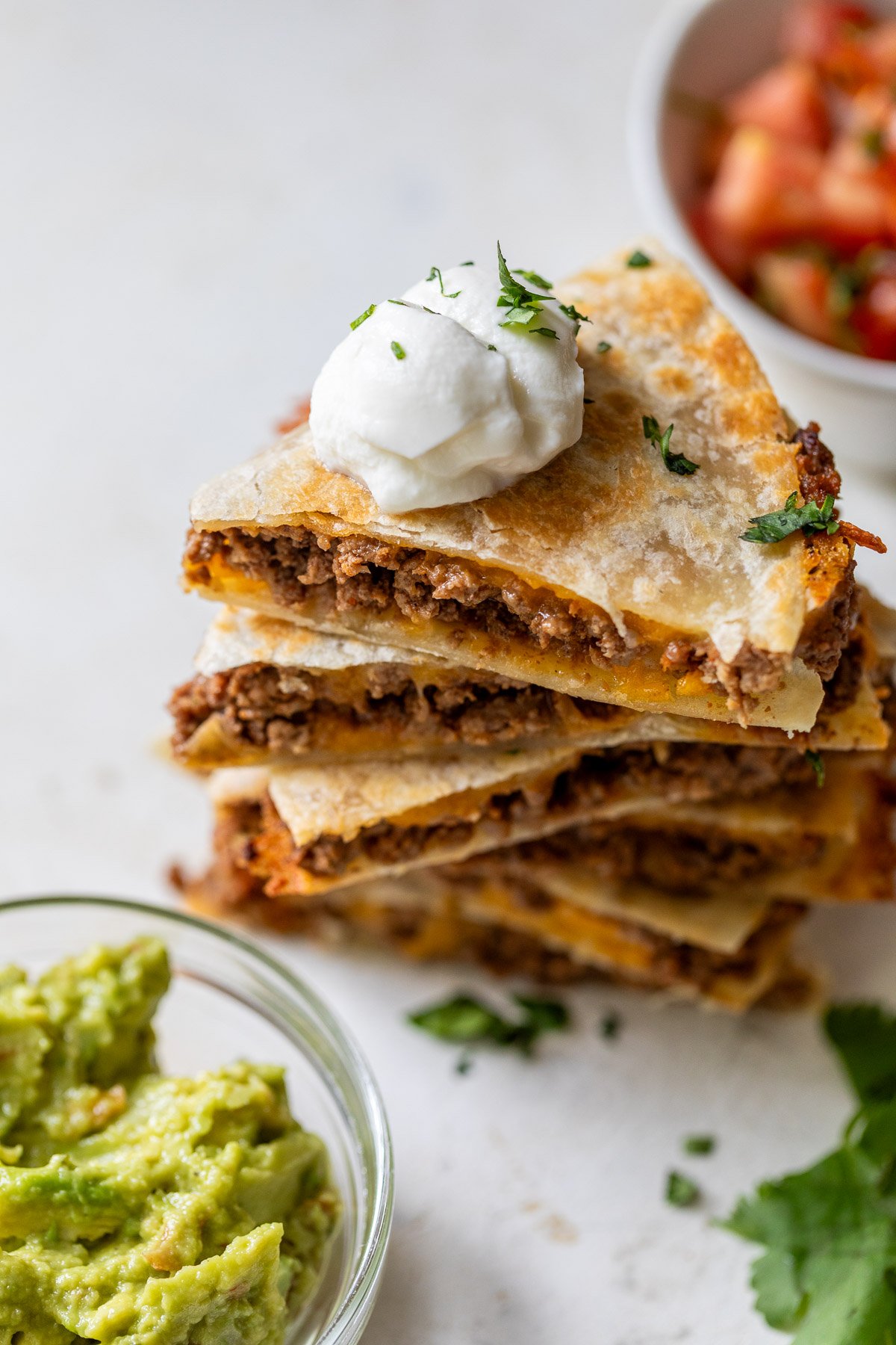 A stack of cheese and beef quesadillas topped with sour cream and cilantro, next to bowls of guacamole and salsa on a white surface.
