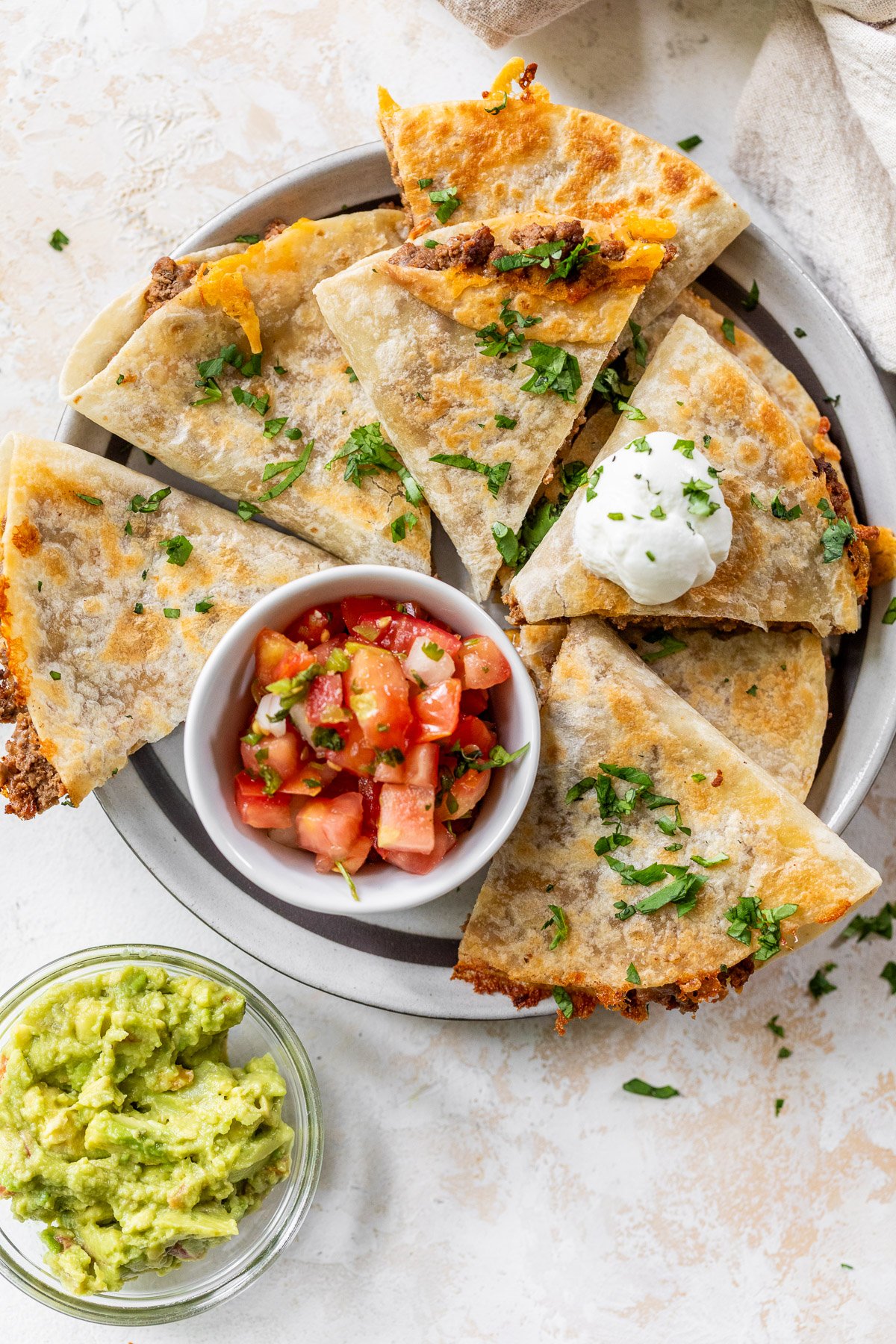 A plate of sliced beef quesadillas topped with chopped cilantro and a dollop of sour cream, served with a small bowl of salsa and a separate bowl of guacamole on the side.