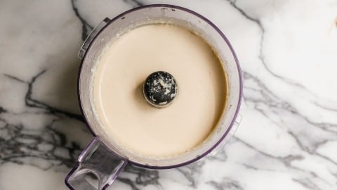 A food processor bowl filled with a smooth, light beige cottage cheese ice cream mixture sits on a white marble countertop.