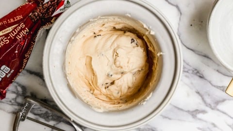 An ice cream maker insert with creamy cottage cheese ice cream sits on a marble countertop, next to a metal ice cream scoop and a partially opened bag of mini semi-sweet chocolate chips.
