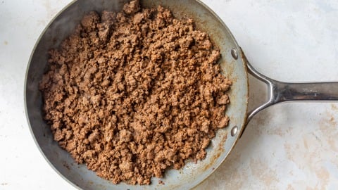 Cooked and browned ground beef in a metal skillet, viewed from above, on a light-colored countertop.