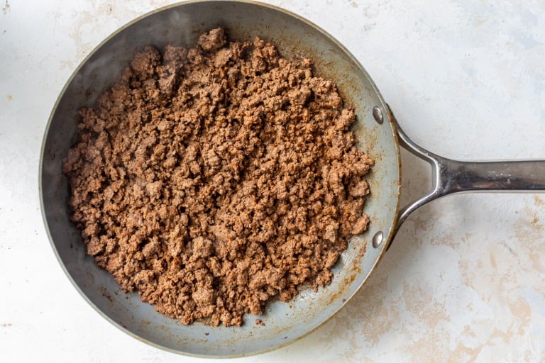 Cooked and browned ground beef in a metal skillet, viewed from above, on a light-colored countertop.